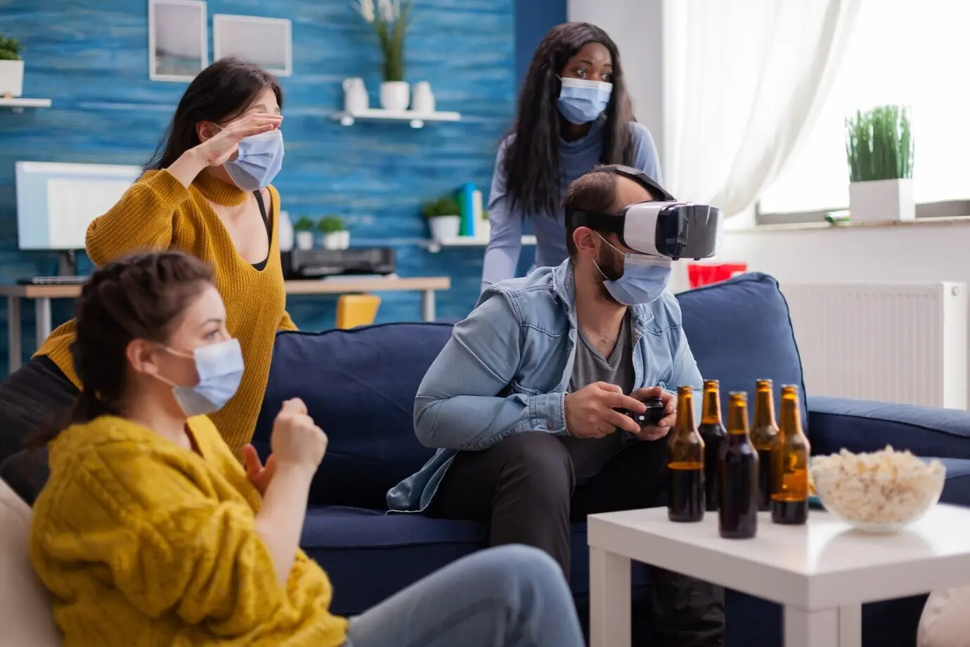 Eine Frau erlebt virtuelle Realität beim Videospielen mit einem VR-Headset und trägt eine Gesichtsmaske, während Freunde aufmunternd jubeln, Abstand halten und ebenfalls Masken tragen, um eine Virusinfektion zu verhindern, Bier.