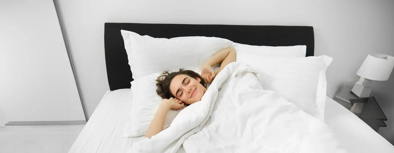 Porträt einer lächelnden, zufriedenen jungen Frau, die beim Aufwachen nach gutem Nachtschlaf liegend die Arme ausstreckt.
